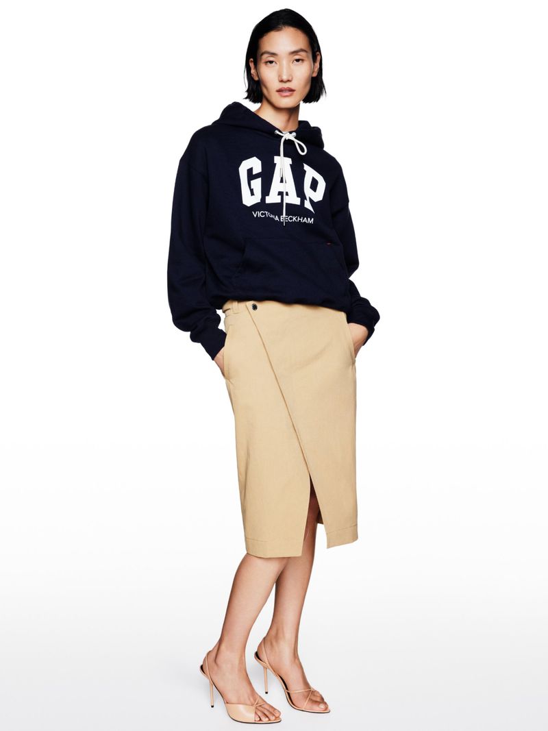 Fashion portrait of a woman in a navy GAP hoodie and khaki midi skirt posing in a studio with beige high-heeled shoes.