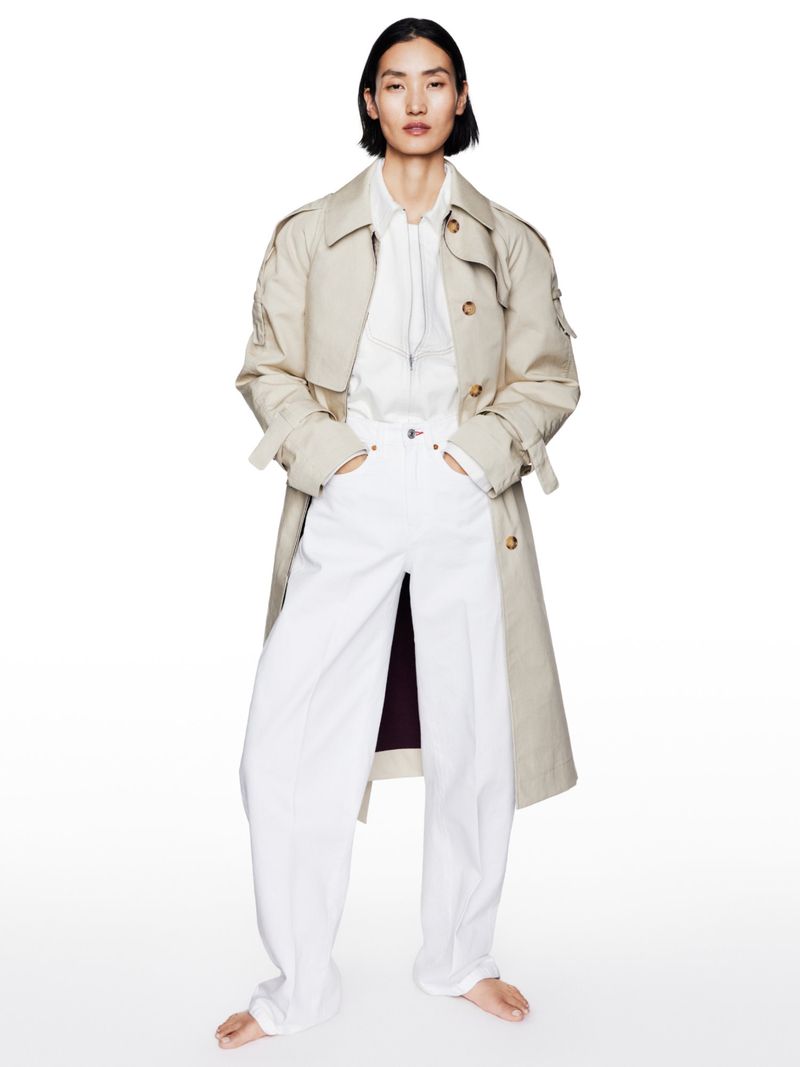 Fashion editorial shot of an adult woman in a beige trench coat, white shirt, and white pants, posing in a photo studio against a white backdrop.