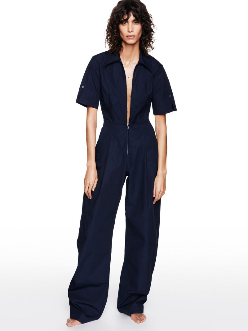 Fashion model posing in a navy blue short-sleeved, wide-legged jumpsuit against a white backdrop in a studio setting, full-length.
