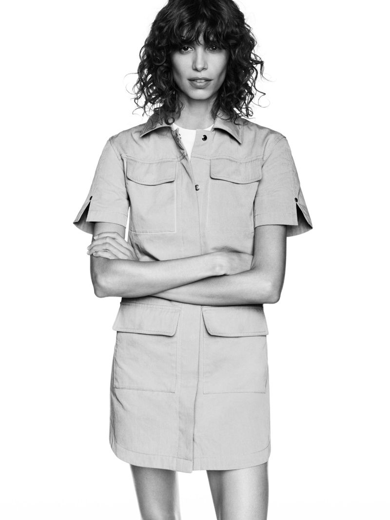 A woman with curly hair wearing a light brown shirt dress poses against a white backdrop in a studio shot.