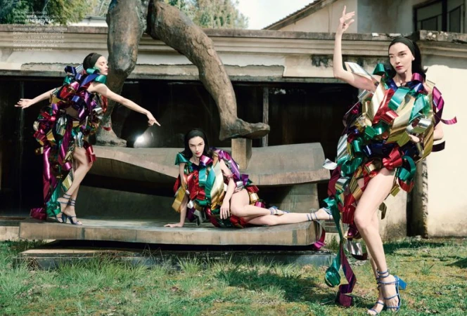 Three women in ribbon dresses pose next to a classical sculpture in an outdoor editorial fashion shot with natural lighting.