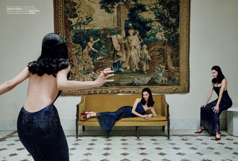 Three women in elegant gowns pose in an opulent indoor setting with a large tapestry, creating a classic fashion editorial look.