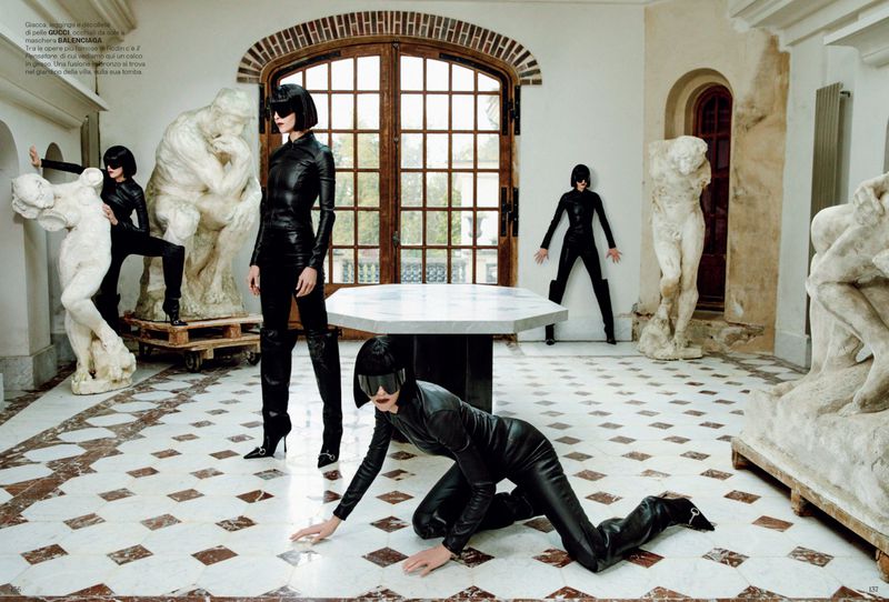 Four women in black catsuits pose with classical sculptures in a white-walled hall, creating a surreal fashion editorial scene with marble floors.