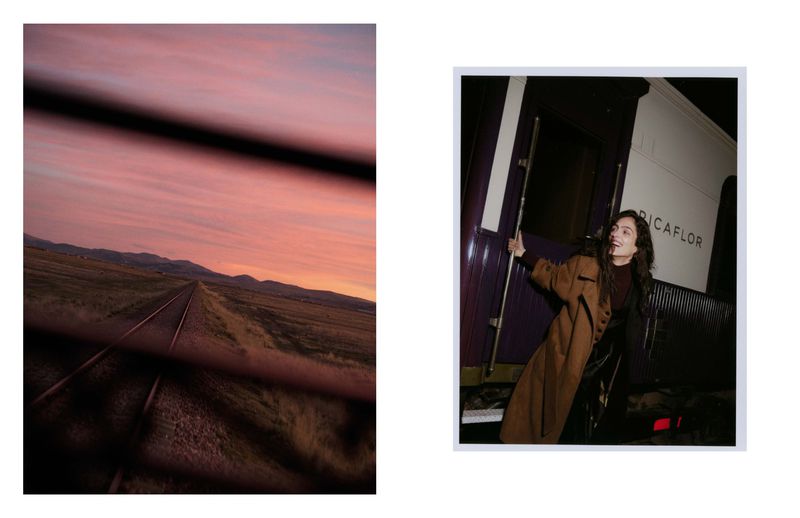 Model poses by train car on tracks extending to horizon, framed by train window bars, at sunset with colorful sky in background.