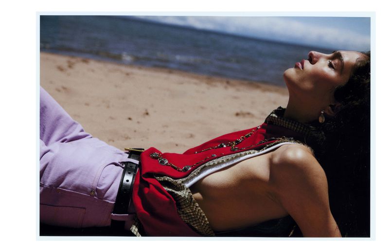 Woman in a red vest and lavender pants lays on a beach with dark hair and a black belt, eyes closed, enjoying the sunlight.