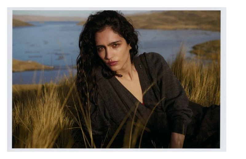 Portrait of a brunette woman with curly hair lying in tall grass with a lake and hills in the background, wearing a grey shawl.