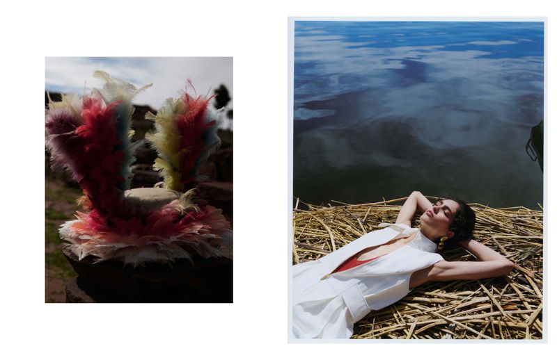 Woman in white dress reclines on woven mat by water with mirrored sky; colorful feather headdress on stone platform outdoors.