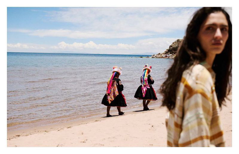 Two young girls in traditional dress walk on a sandy beach, with the ocean and a woman in the foreground, creating a vibrant scene.