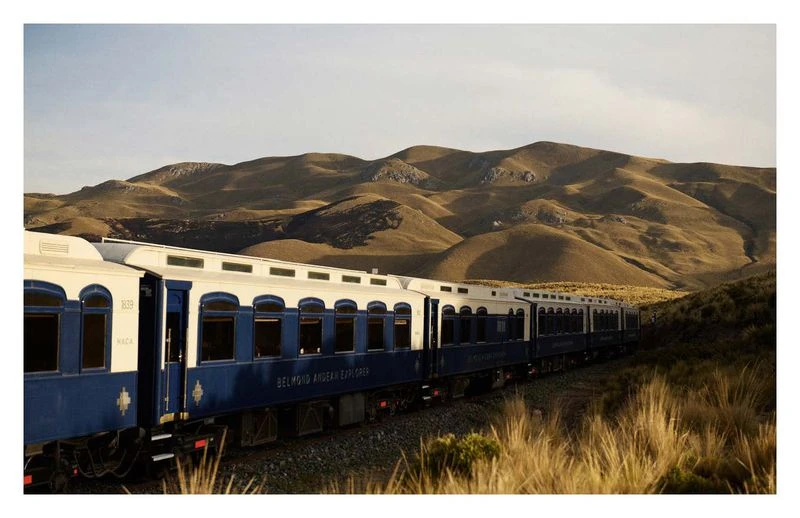 The Belmond Andean Explorer train travels through a valley with golden grasses against a backdrop of rolling, bare mountains under a blue sky.