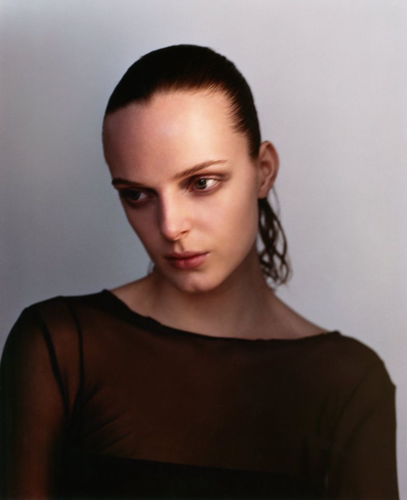 Close-up portrait of a young adult woman with slicked-back dark hair, wearing a sheer dark top, gazing downward in a softly lit photo studio.
