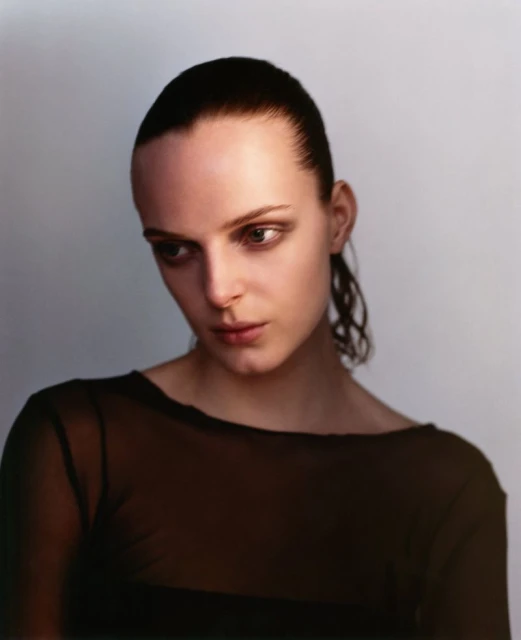 Close-up portrait of a young adult woman with slicked-back dark hair, wearing a sheer dark top, gazing downward in a softly lit photo studio.