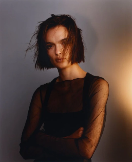 Portrait of a brunette adult woman with arms crossed, wearing a sheer black top. The lighting is warm, with a gray background.