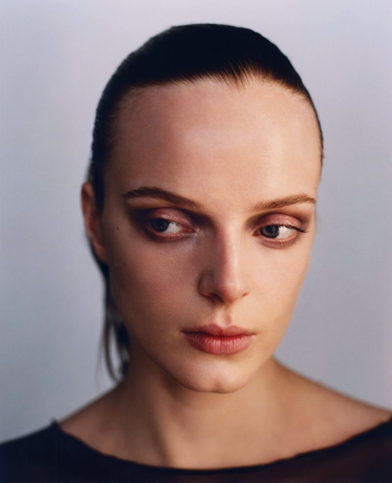 Close-up portrait of an adult woman with slicked-back dark hair, wearing a sheer black top against a light gray background.