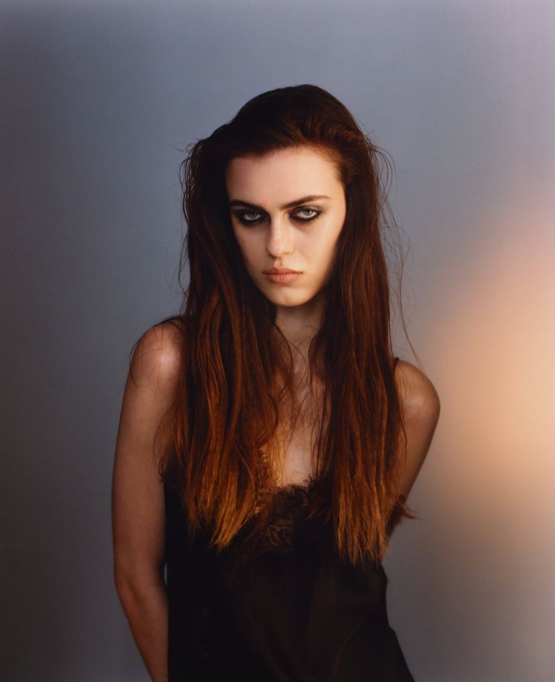 Portrait of a woman with long dark hair and dramatic dark eye makeup wearing a black slip top against a neutral gradient studio background.