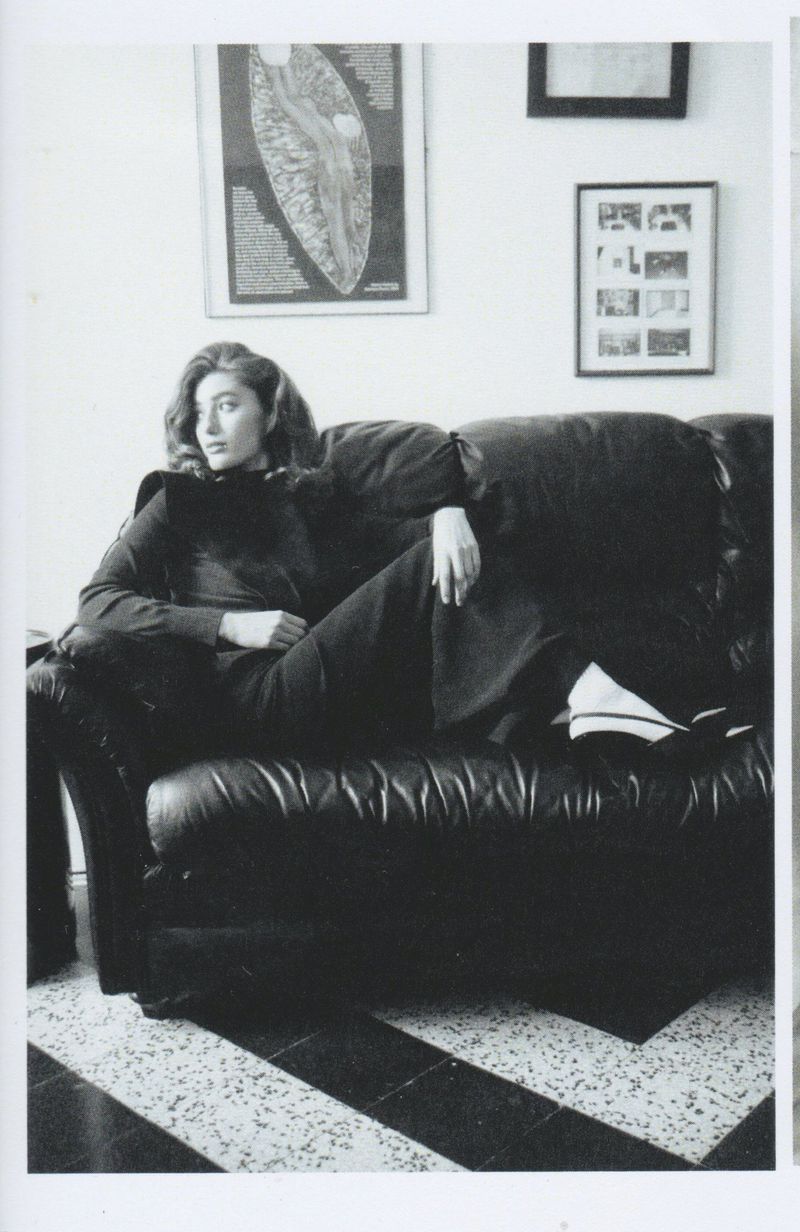 Black-and-white portrait of a woman in a dark outfit reclining on a black leather couch with patterned flooring and framed artwork on the wall.