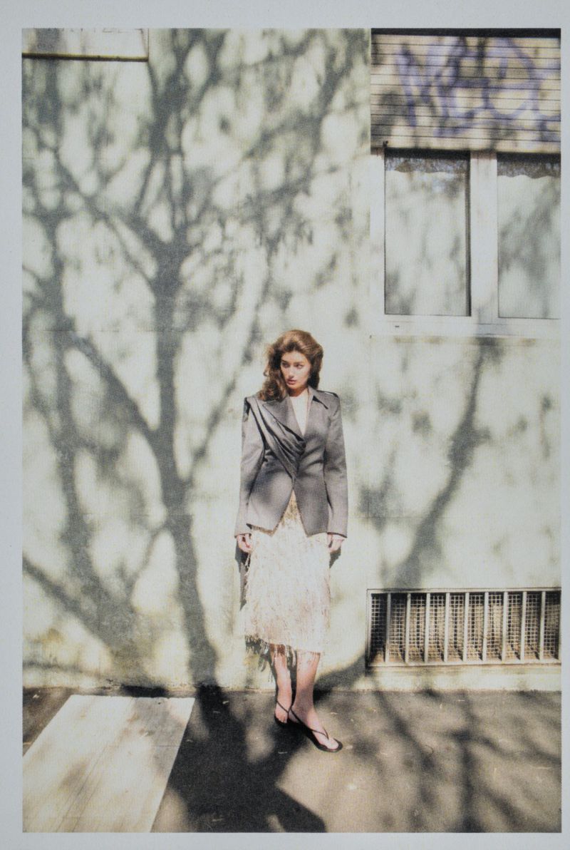 Woman in blazer and dress stands against a shadowed wall next to a window with bars, creating an artistic portrait in natural light.