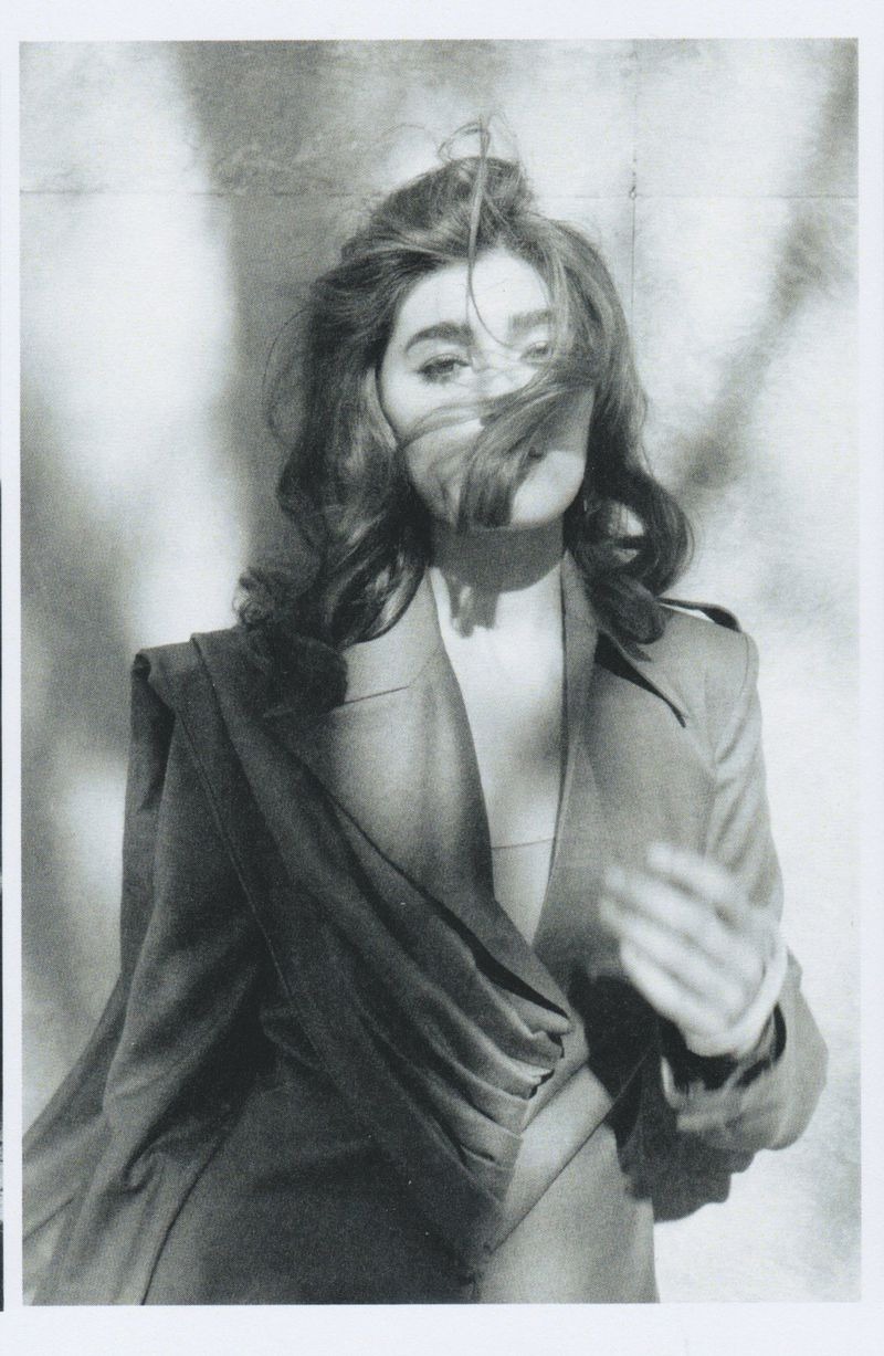Monochrome portrait of a woman with wavy brown hair wearing a draped blazer against a blurred patterned backdrop, looking at the camera.