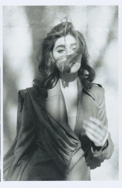 Monochrome portrait of a woman with wavy brown hair wearing a draped blazer against a blurred patterned backdrop, looking at the camera.