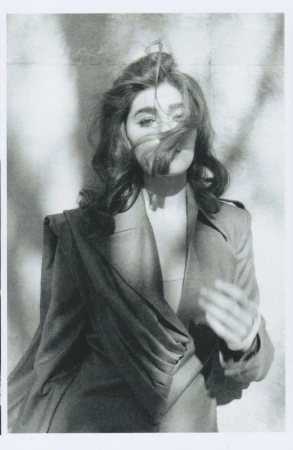 Monochrome portrait of a woman with wavy brown hair wearing a draped blazer against a blurred patterned backdrop, looking at the camera.