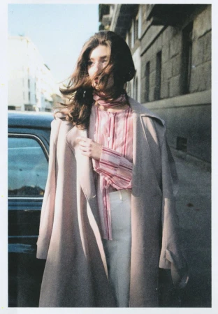 Brunette woman in tan coat and pink blouse walking on an urban street near a parked car on a sunny day with wind blowing her hair.