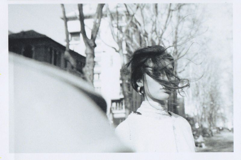 A young woman with her hair blowing in the wind stands on an urban street, captured in black and white, trees in the background.