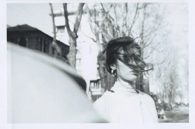 A young woman with her hair blowing in the wind stands on an urban street, captured in black and white, trees in the background.