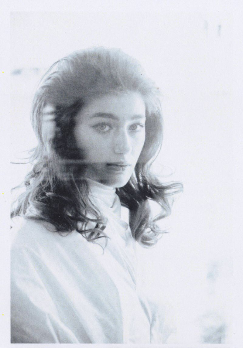 Monochrome portrait of a young woman with voluminous dark hair wearing a light-colored top, looking directly at the camera.