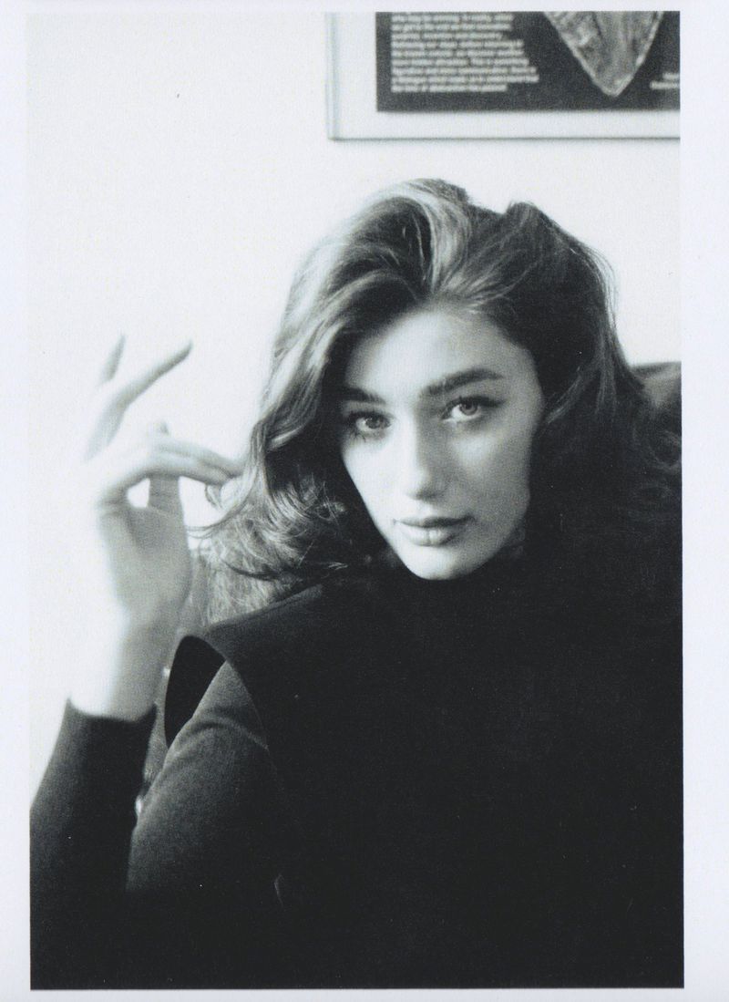Portrait of a woman with dark wavy hair wearing a black top, with a framed picture visible on the white wall behind her.