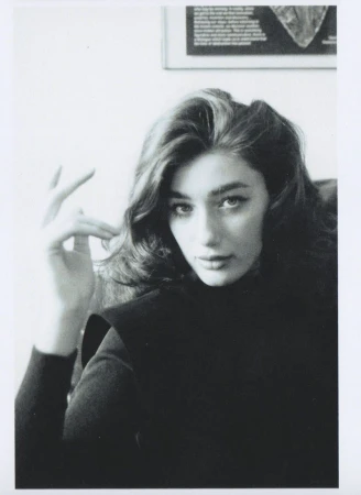 Portrait of a woman with dark wavy hair wearing a black top, with a framed picture visible on the white wall behind her.