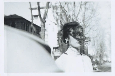 Black and white polaroid photograph of a woman with windblown hair outdoors with urban trees and buildings in the background.