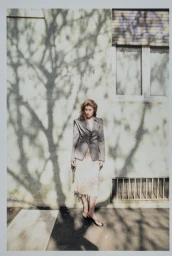 Model standing outdoors against a pale wall with tree branch shadows, wearing a structured grey jacket and patterned skirt.