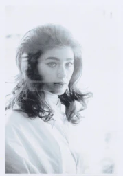 Portrait of a woman with dark voluminous hair and winged eyeliner wearing a white top against a bright white background