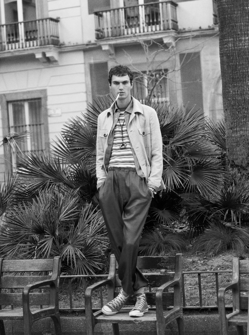 Monochrome fashion portrait of a man standing on a park bench outdoors against palm plants and classic building architecture.