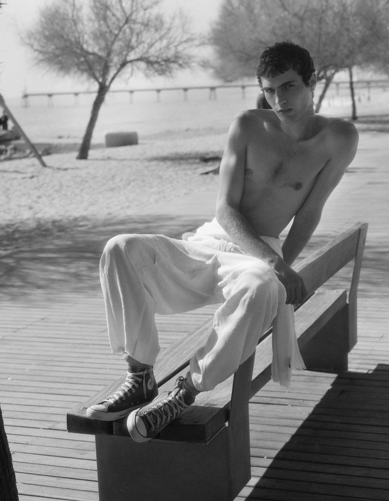 Black and white photo of a young man with no shirt on sitting on a bench near a beach, wearing white pants and converse sneakers.