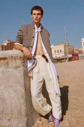 Male model leaning against a concrete wall outdoors wearing loose white trousers and an open jacket under a clear sky.