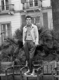 Monochrome fashion portrait of a man standing on a park bench outdoors against palm plants and classic building architecture.