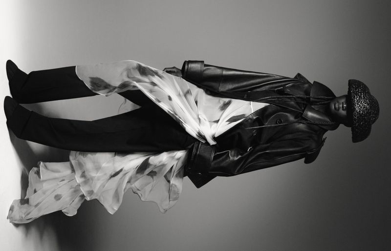 Black and white fashion shot of a model in a leather coat and hat, accessorized with a patterned sheer fabric, in a studio setting.