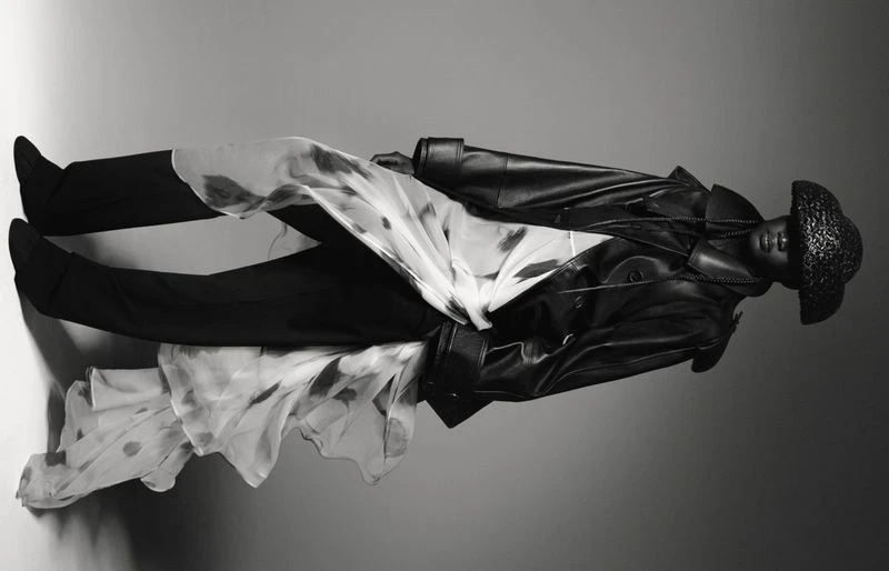 Black and white fashion shot of a model in a leather coat and hat, accessorized with a patterned sheer fabric, in a studio setting.