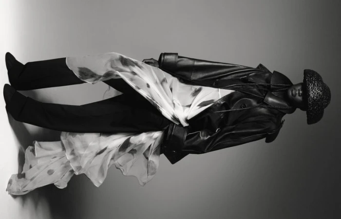 Black and white fashion shot of a model in a leather coat and hat, accessorized with a patterned sheer fabric, in a studio setting.
