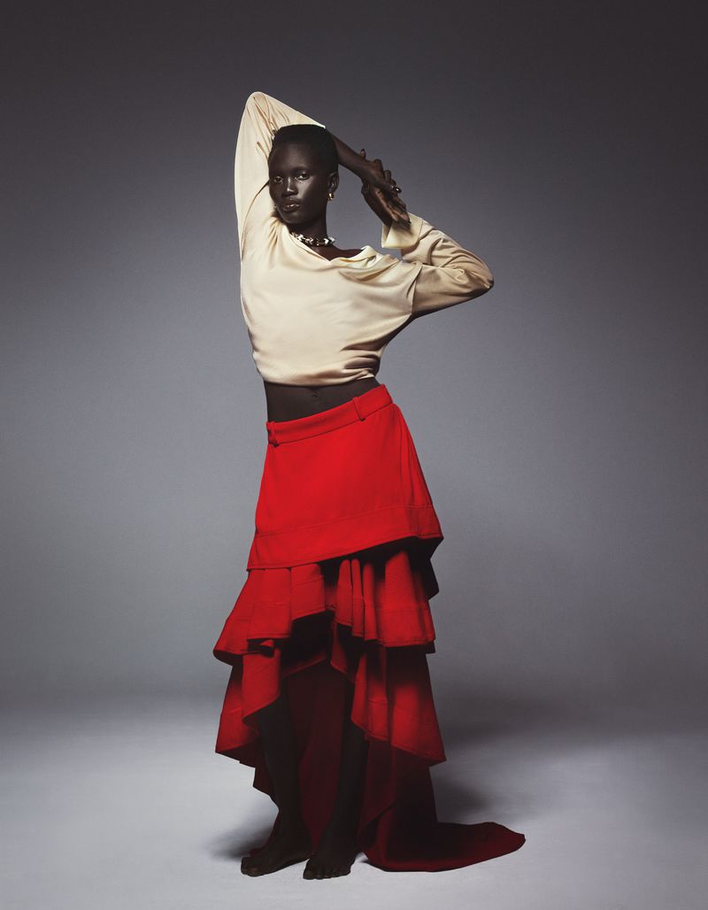 Elegant dark-skinned model poses in a flowing red skirt and cream blouse against a grey backdrop in a studio portrait with soft lighting.