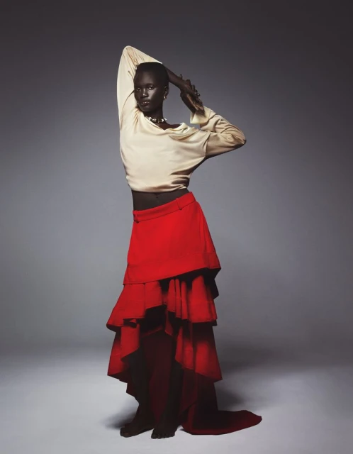 Elegant dark-skinned model poses in a flowing red skirt and cream blouse against a grey backdrop in a studio portrait with soft lighting.