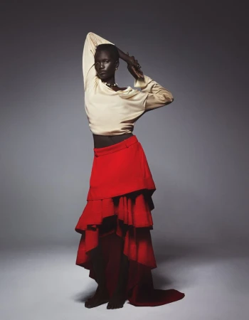Elegant dark-skinned model poses in a flowing red skirt and cream blouse against a grey backdrop in a studio portrait with soft lighting.