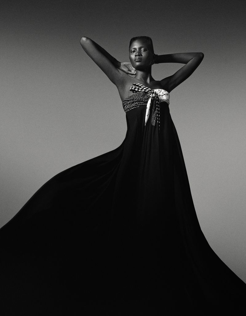 Elegant dark-skinned woman in strapless black gown with flowing train poses hands behind head in a studio fashion shot against gray backdrop.