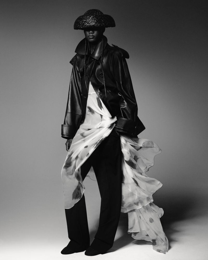 Fashion editorial shot of a model wearing a black leather jacket, matching trousers, straw hat, and patterned white dress in a studio.