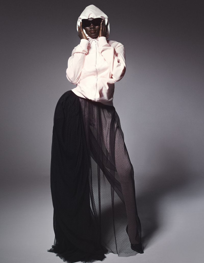 A person in a light pink hooded sweatshirt and a long black mesh skirt poses in a studio with hands near their face.