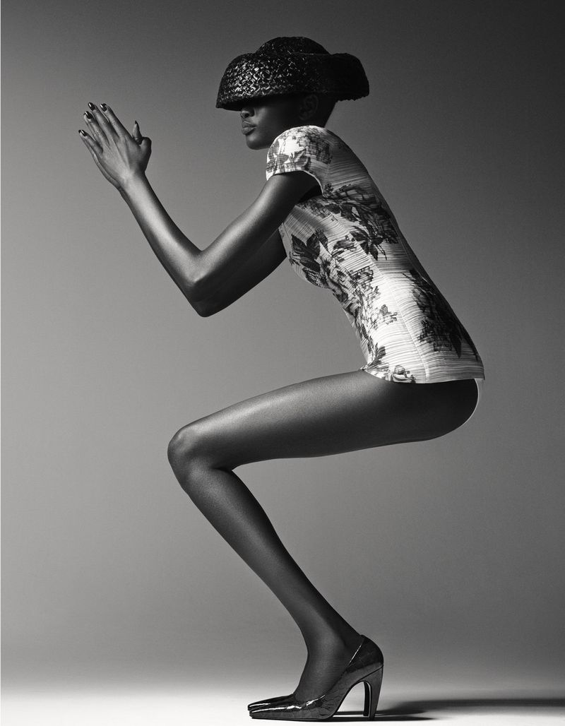 A fashion-style studio shot features an African American woman wearing a floral blouse, hat, and high heels, in a dynamic crouching pose.