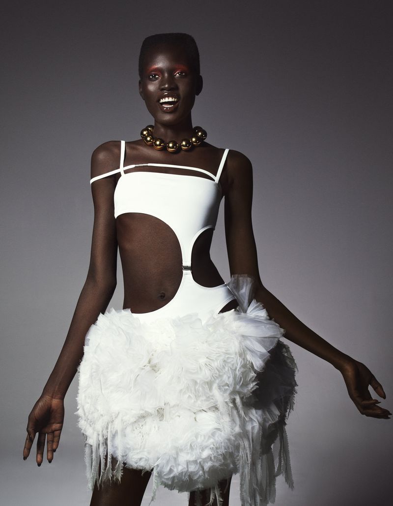 Woman in a white, avant-garde dress with feathered skirt, gold necklace, and dramatic makeup, posing against a neutral gray background.