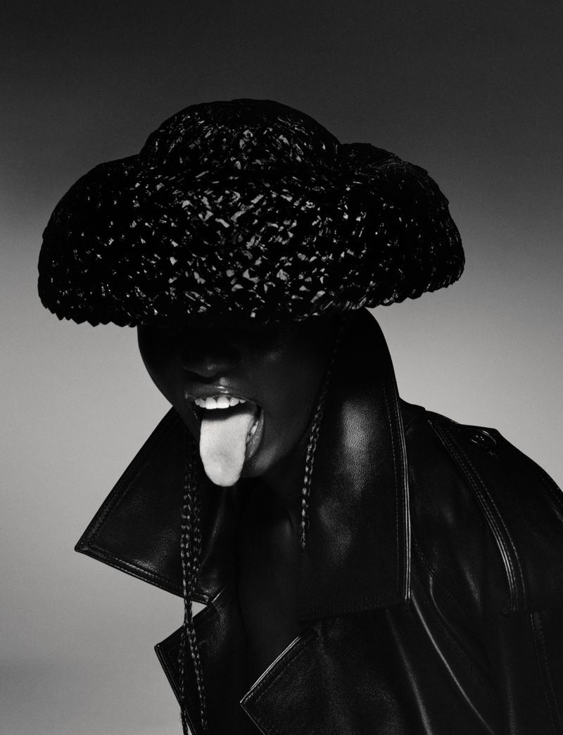 Close-up portrait of a person with dark skin wearing a large black woven hat and a black leather jacket, sticking their tongue out.