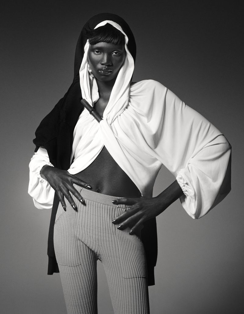 Black and white fashion portrait of a dark-skinned woman in a hooded sweater and ribbed pants, posing in a photo studio.