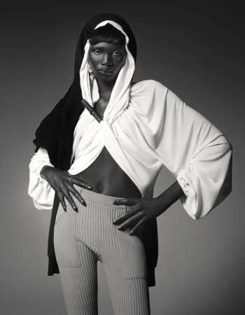Black and white fashion portrait of a dark-skinned woman in a hooded sweater and ribbed pants, posing in a photo studio.
